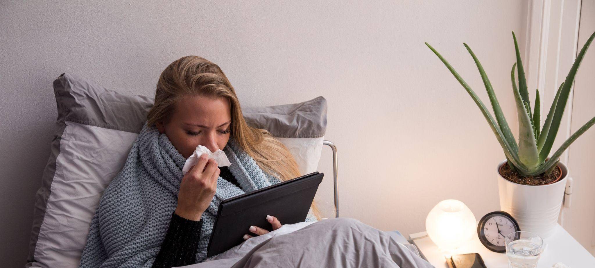 Eine Frau liegt krank im Bett und guckt auf ihren Tablet-Computer